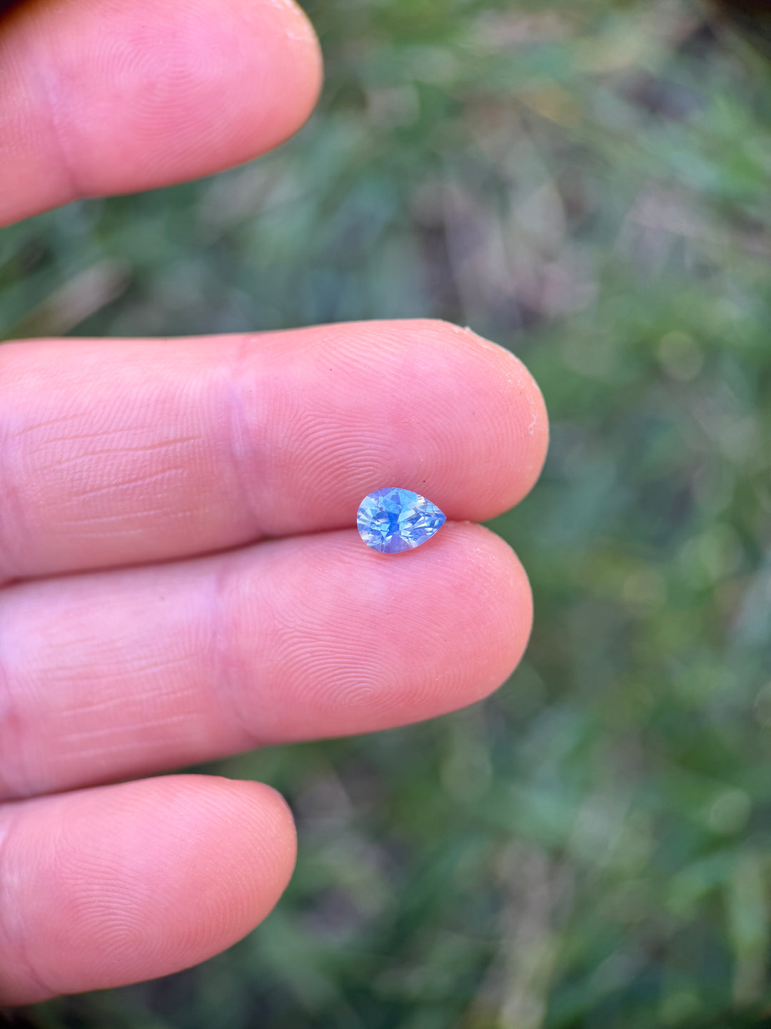 Loose 1.11 carat opalescent cornflower blue Nigerian sapphire resting on fingertips, highlighting the brilliant pear cut and natural transparency.