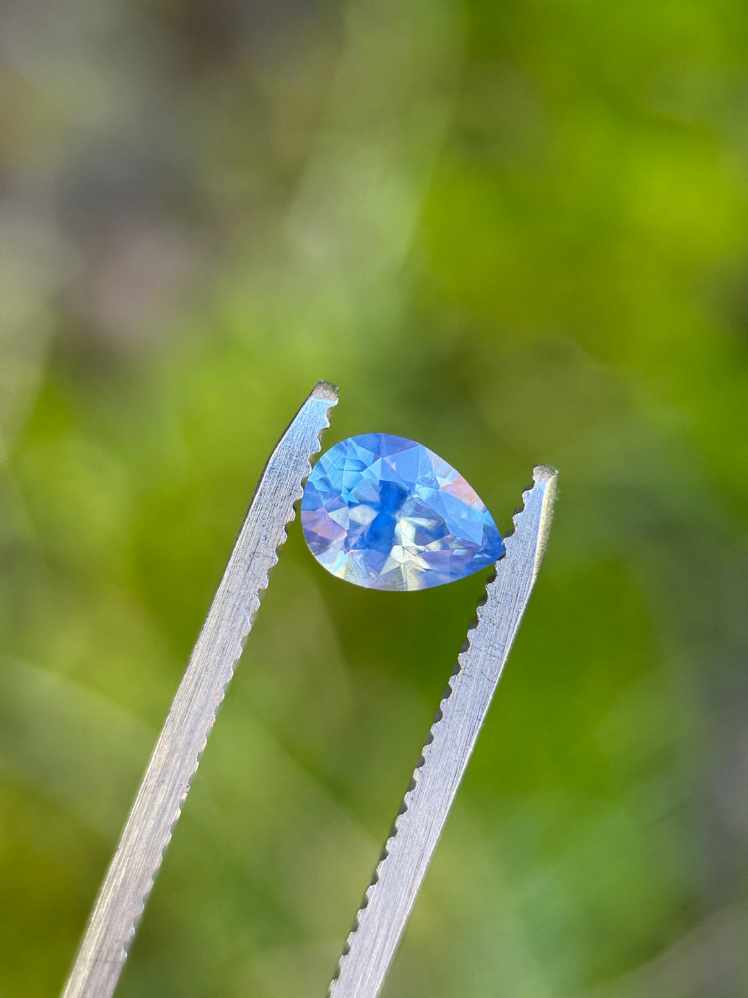 Loose 1.11 carat opalescent cornflower blue sapphire from Nigeria held in tweezers, showing the precision brilliant pear cut and lively facet reflections.