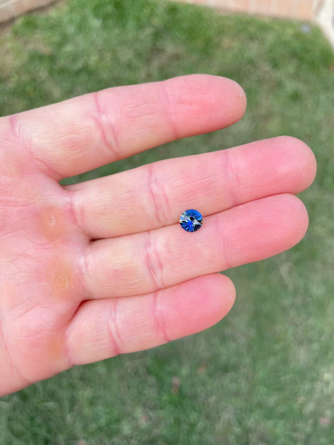 A 1.11-carat round brilliant parti sapphire with a vibrant mix of blue and yellow hues, displayed on a hand in natural daylight.