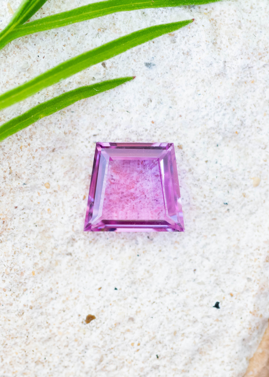Hot pink 1.12ct portrait cut sapphire resting on a textured white surface with small green leaves coming into frame.