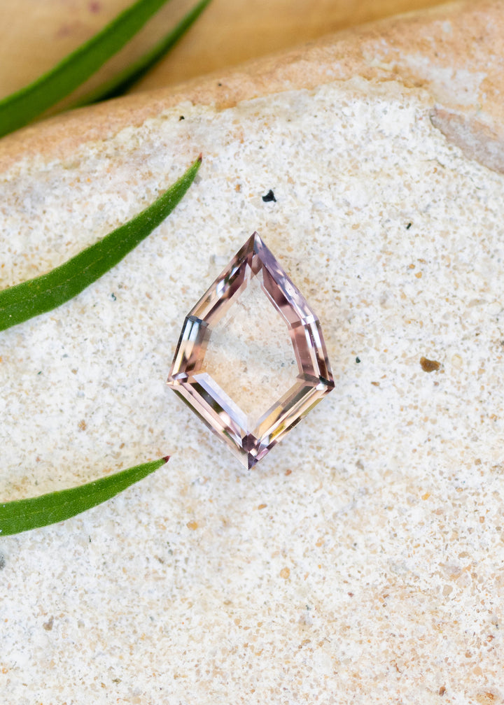 1.12ct peach and blue parti portrait cut sapphire resting on white sandy surface with leaves resting on the side.