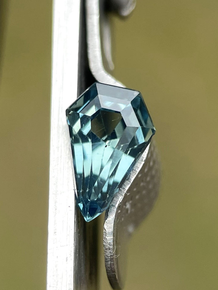 Close-up of a 1.13-carat opalescent denim blue sapphire, cut in a precise step cut shield shape, held by metal tweezers to show its luster. Originating from Madagascar, this no-heat treated gem showcases exceptional clarity and a unique geometric design.