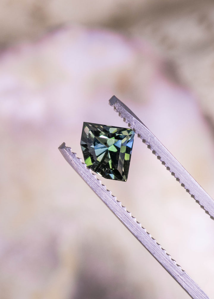 Loose forest green tanzanite held in tweezers, showing the custom geometric fan mix cut and natural color shift under direct light.