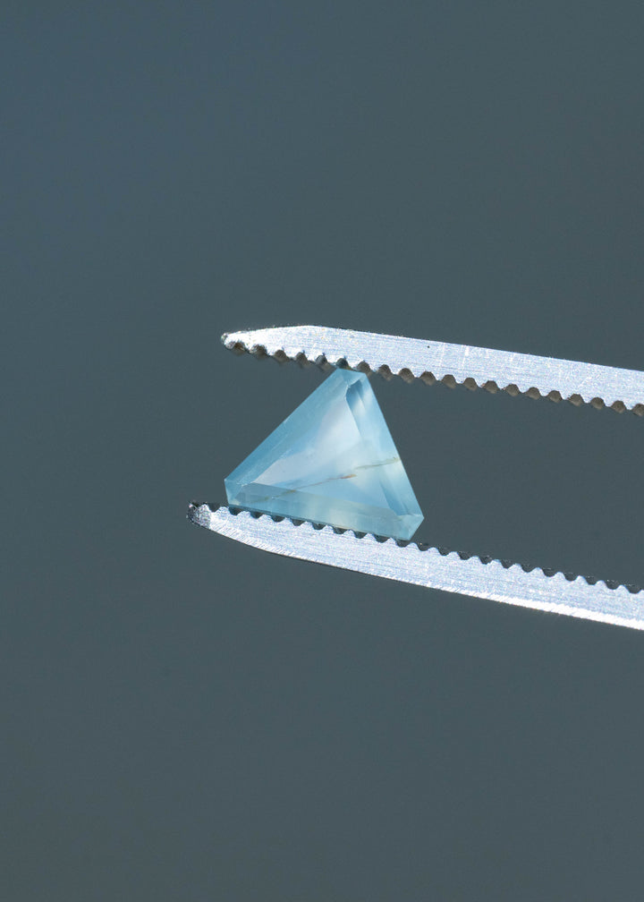 Small, pale blue triangular sapphire held between silver tweezers against a dark gray background. Its translucent, misty interior contains a fine brownish line, with lighter blue facets around the edges.