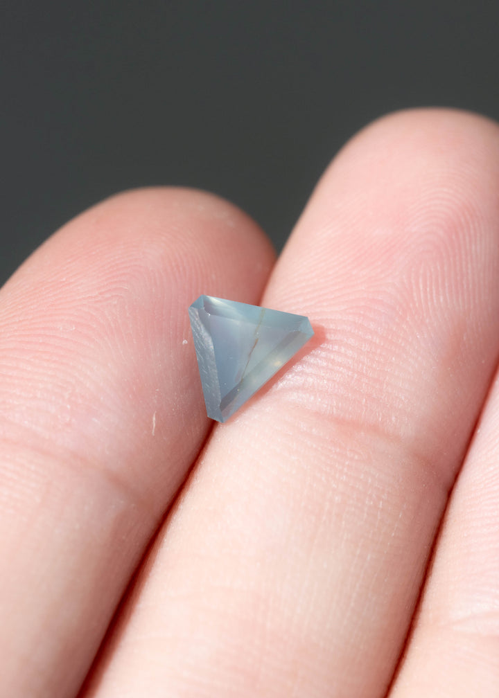 Pale blue-gray triangular sapphire resting across two fingertips. Its broad, softly clouded center contains a thin brownish inclusion, while angular edge facets define its three-sided shape.