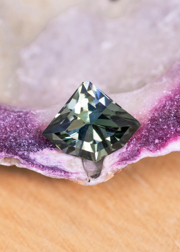 Loose teal, yellow, and green tanzanite from Merelani Hills, Tanzania shown face-up on a mineral surface, displaying the geometric shield mix cut and sharp facet structure.