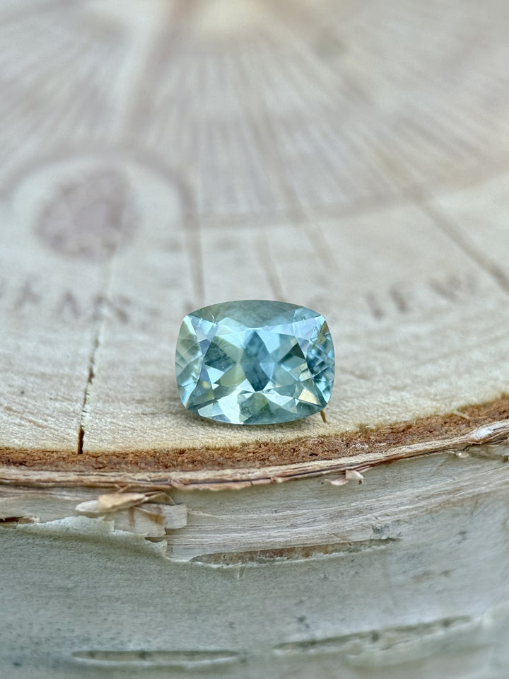 1.15 carat teal blue-green sapphire from El Dorado Bar, Montana resting on wood log, custom elongated cushion cut, untreated natural stone