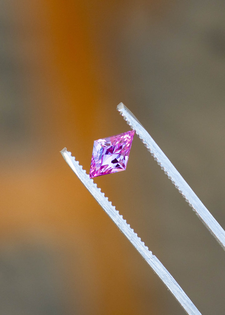 1.15ct hot pink custom kite rhombus cut sapphire held in gem tweezers showing off natural vibrant hue under natural lighting.