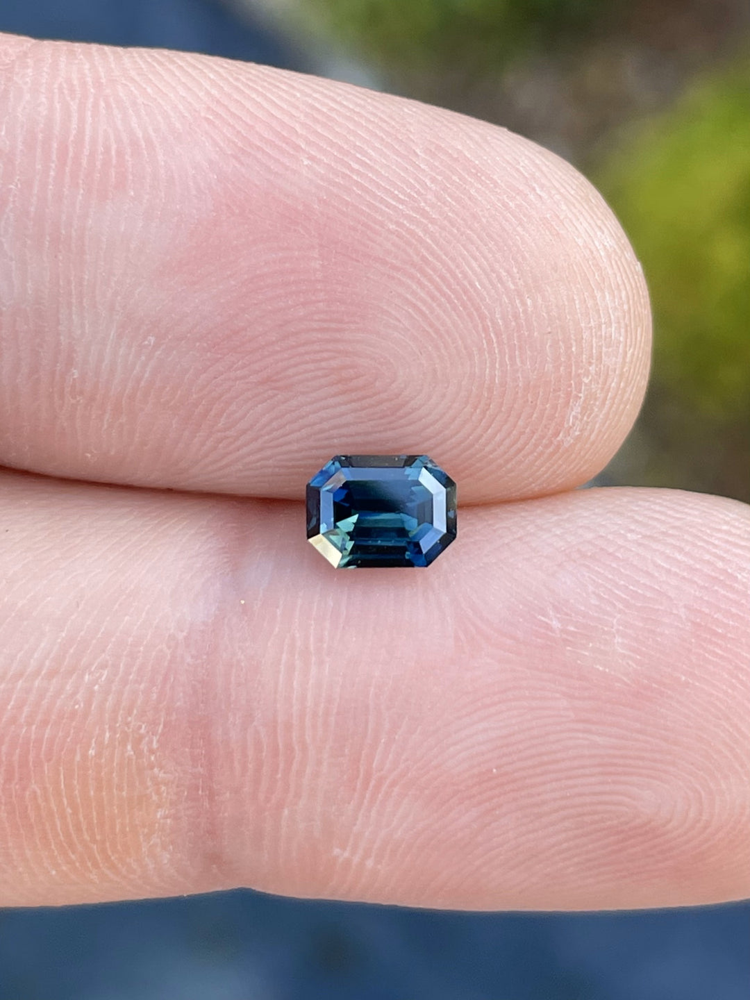 A stunning 1.16-carat green-blue teal sapphire in an octagonal step cut, held between fingers to showcase its brilliance and color shift. Origin: Gombe, Nigeria.