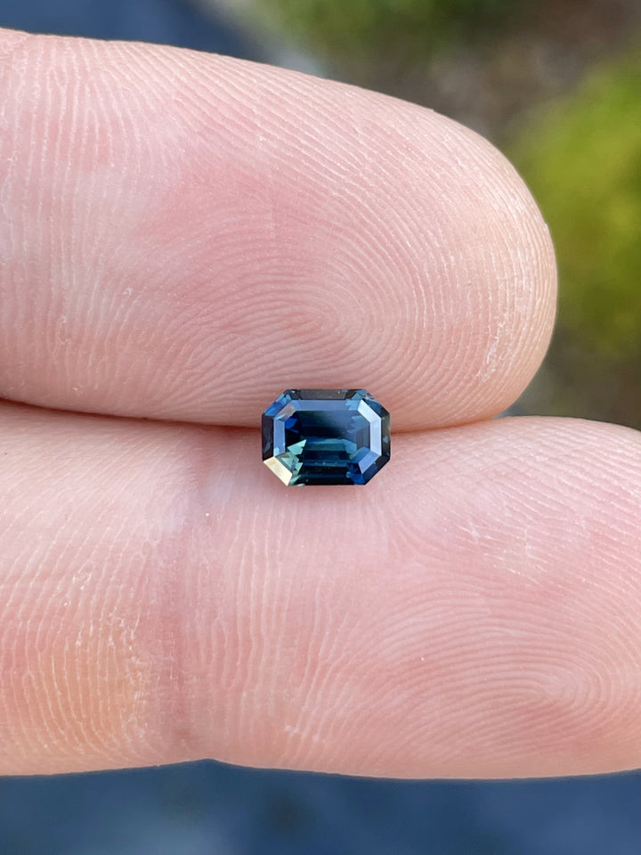 A stunning 1.16-carat green-blue teal sapphire in an octagonal step cut, held between fingers to showcase its brilliance and color shift. Origin: Gombe, Nigeria.