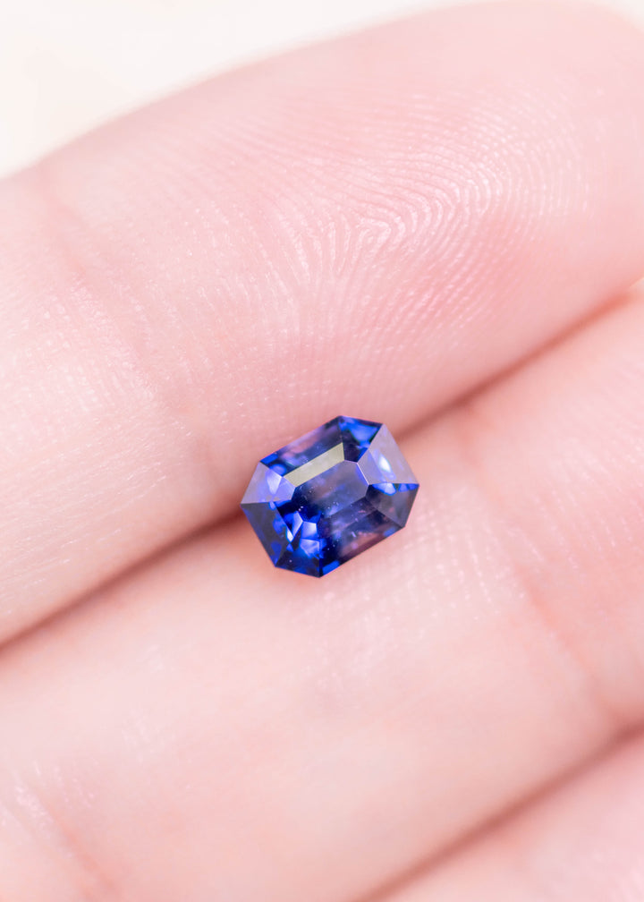 Loose blue bi-color sapphire from Sri Lanka shown resting on a fingertip displaying the emerald cut and natural contrast between deep blue and colorless zones.