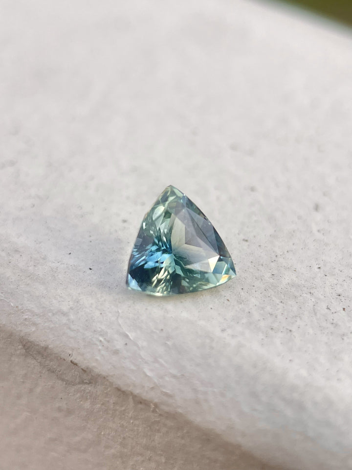 Close-up image of a 1.17-carat bi-color sapphire in a trillion brilliant mixed cut, displaying vibrant blue-green and yellow hues. The gem, originating from Madagascar, is untreated and positioned to highlight its unique color transitions and facets