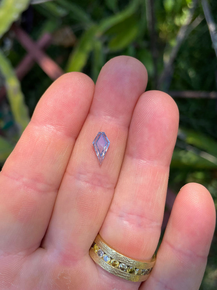 A 1.18-carat custom step cut portrait kite sapphire in a pastel cornflower blue color, held delicately between fingers, with a gold and diamond ring visible, showcasing the gemstone's clarity and unique cut.