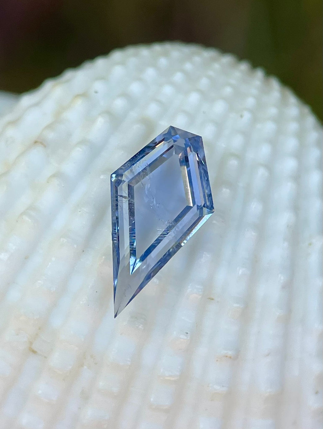 Close-up of a 1.18-carat custom step cut portrait kite sapphire in a pastel cornflower blue color, resting on a textured white surface, highlighting its intricate facets and delicate hue.