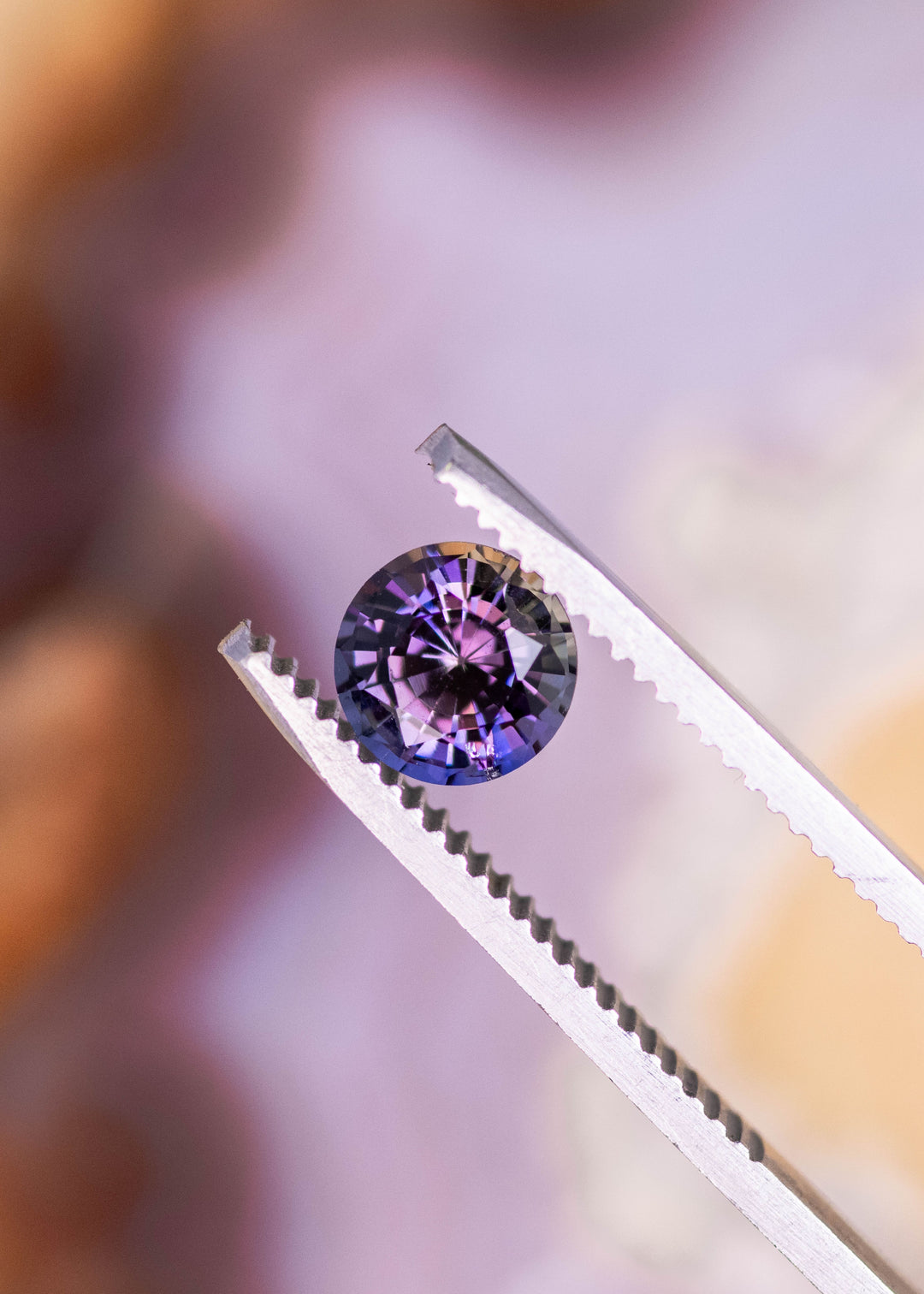 1.19 carat orange purple Tanzanite round brilliant held in tweezers, untreated Merelani Hills Arusha Tanzania gemstone displaying sharp facet reflections, trichroic color and exceptional brilliance for jewelry design