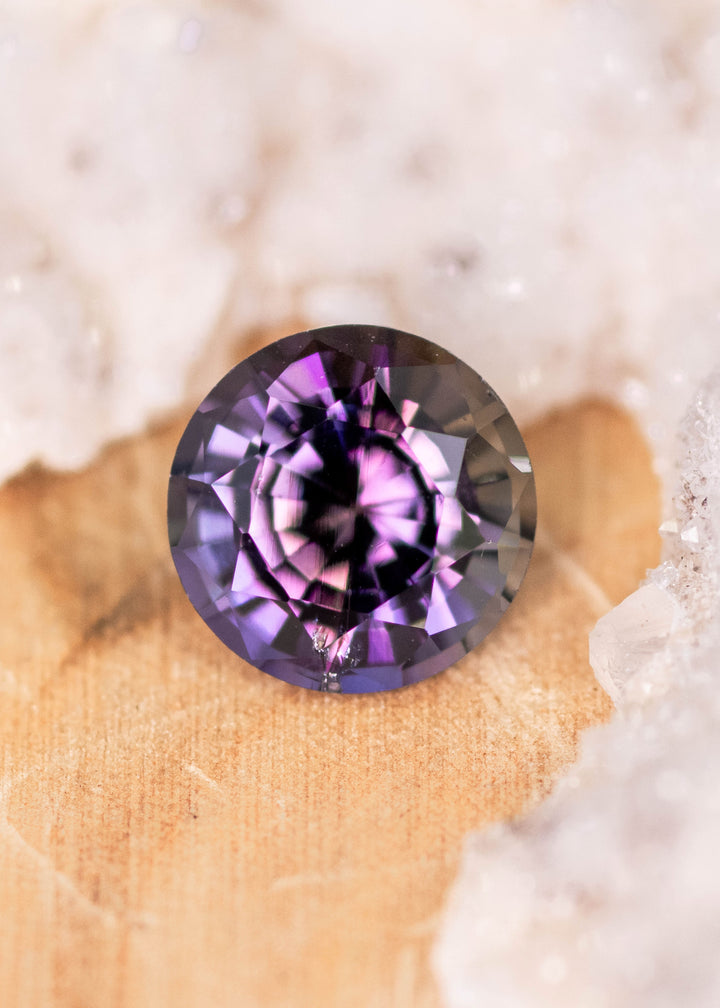 1.19 carat orange purple Tanzanite mix round brilliant placed on display surface, untreated gemstone from Tanzania highlighting precision cut symmetry, vibrant bi color tones and natural brilliance for bespoke jewelry