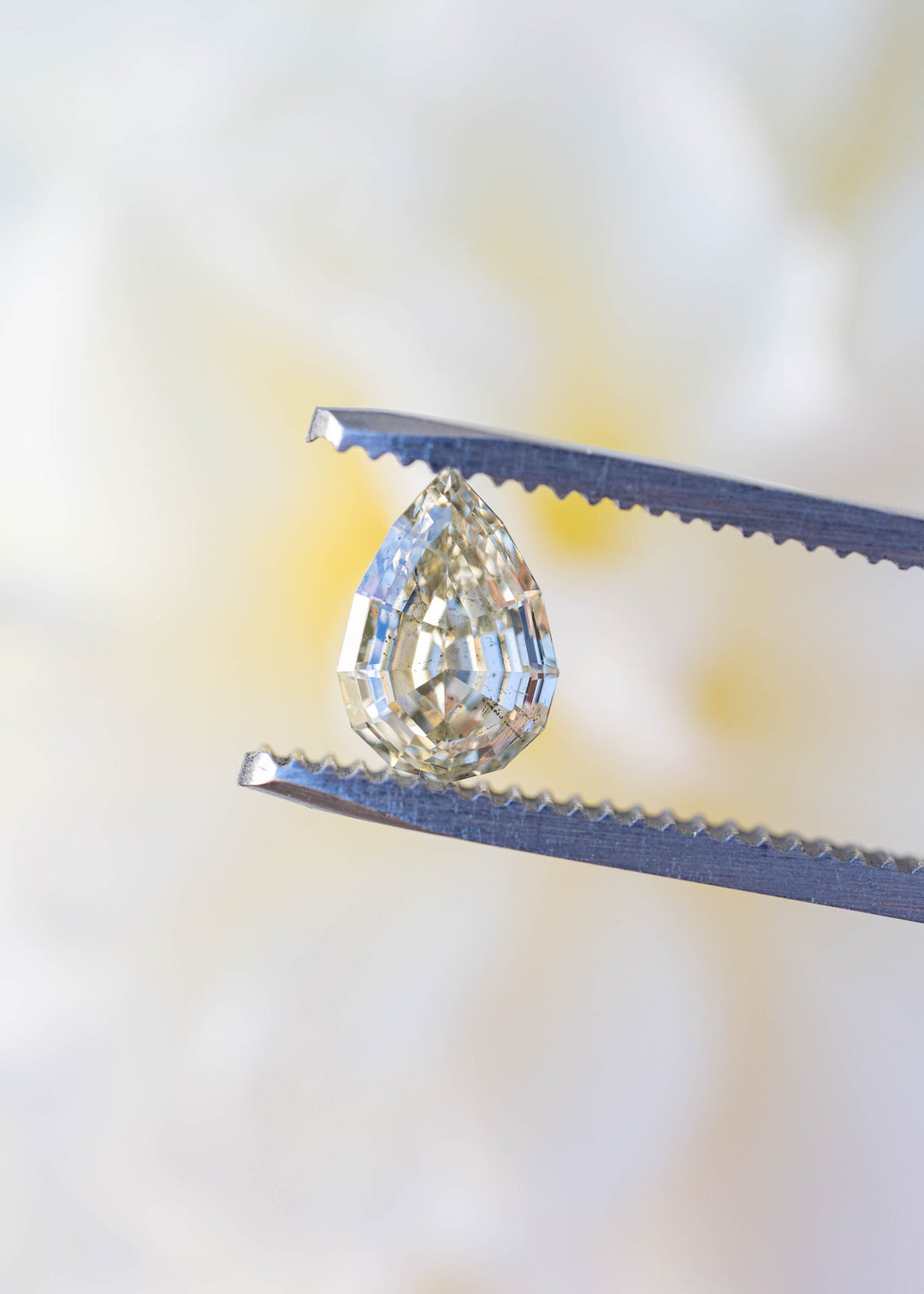 1.20 carat Light Yellow Geo Pear diamond held in jeweler's tweezers against a soft floral background, highlighting its precision step cut and elongated pear shape.