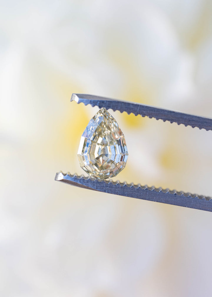 1.20 carat Light Yellow Geo Pear diamond held in jeweler's tweezers against a soft floral background, highlighting its precision step cut and elongated pear shape.