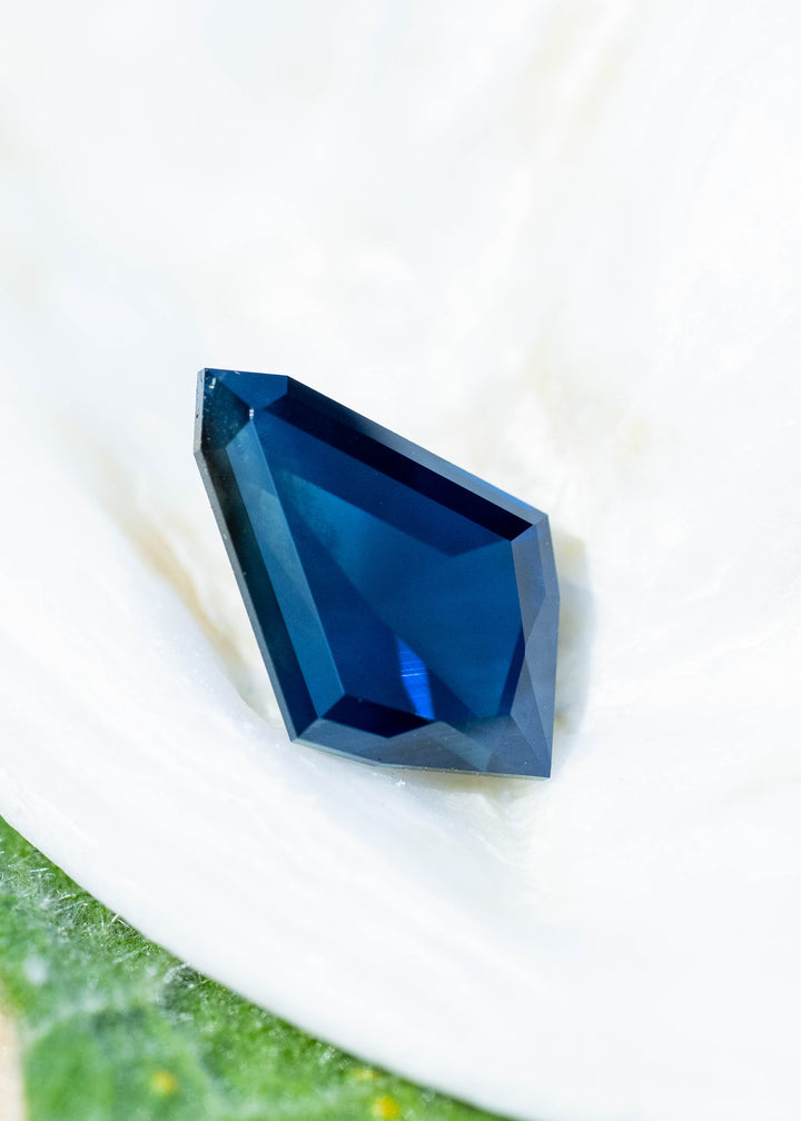 Deep blue, asymmetrical kite-shaped sapphire resting on a white flower petal. Broad geometric facets reveal cobalt-blue reflections within a darker navy center, framed by crisp, angular edges.