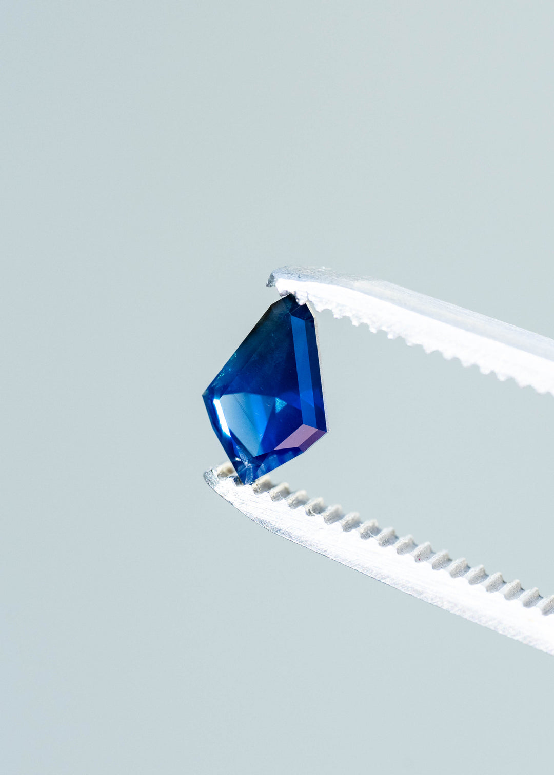 Angular deep blue sapphire held between silver tweezers against a pale blue-gray background. Bright cobalt reflections appear across its lower facets, contrasting with its darker blue center.