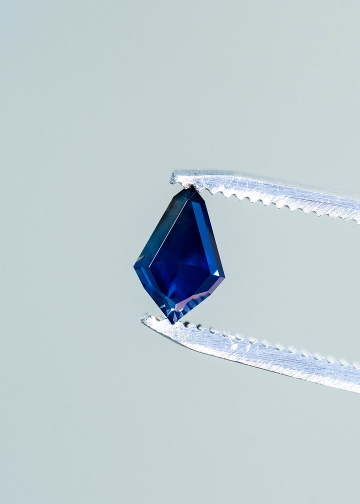 Deep navy-blue kite-shaped sapphire held vertically in silver tweezers. Its broad center appears dark blue, with brighter cobalt reflections near the lower point and a narrow border of polished geometric facets.