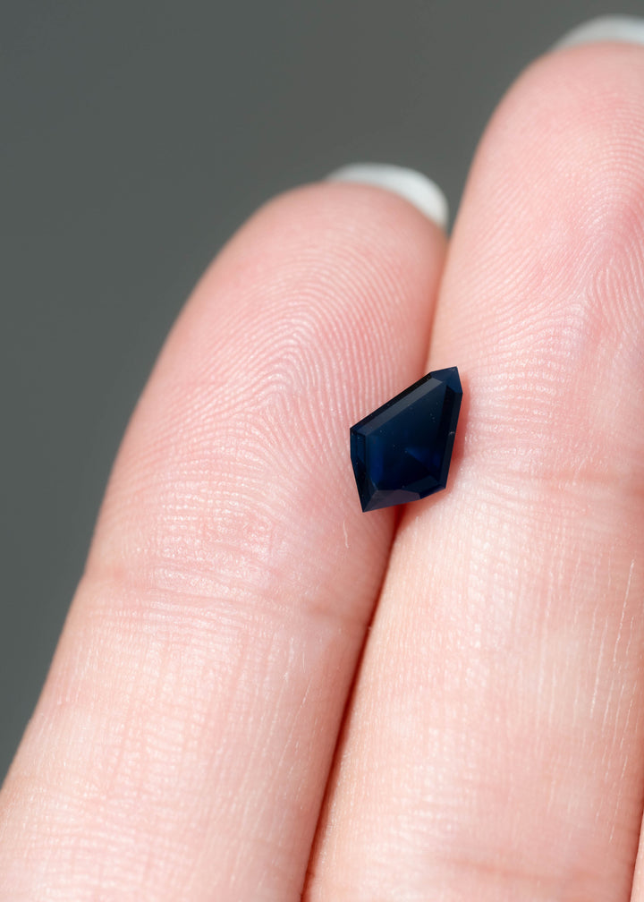 Small, dark blue kite-shaped sapphire resting between two fingertips. Its asymmetrical outline has sharply angled sides, a pointed upper corner and broad facets that appear nearly black-blue in this lighting.