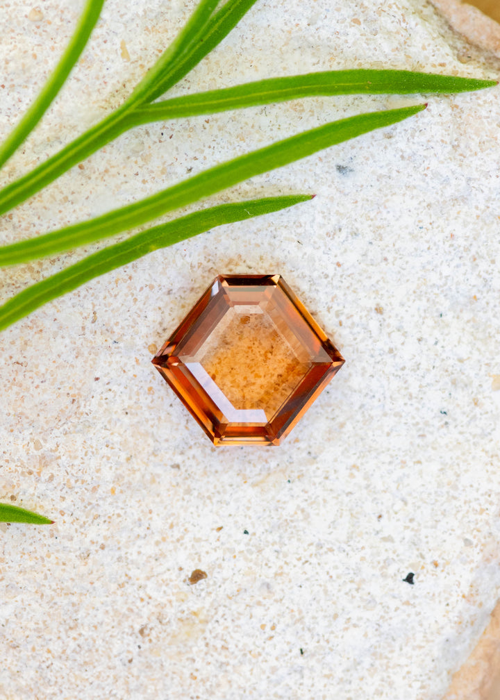 Hexagonal natural orange parti portrait cut sapphire on a sandy surface with green leaves