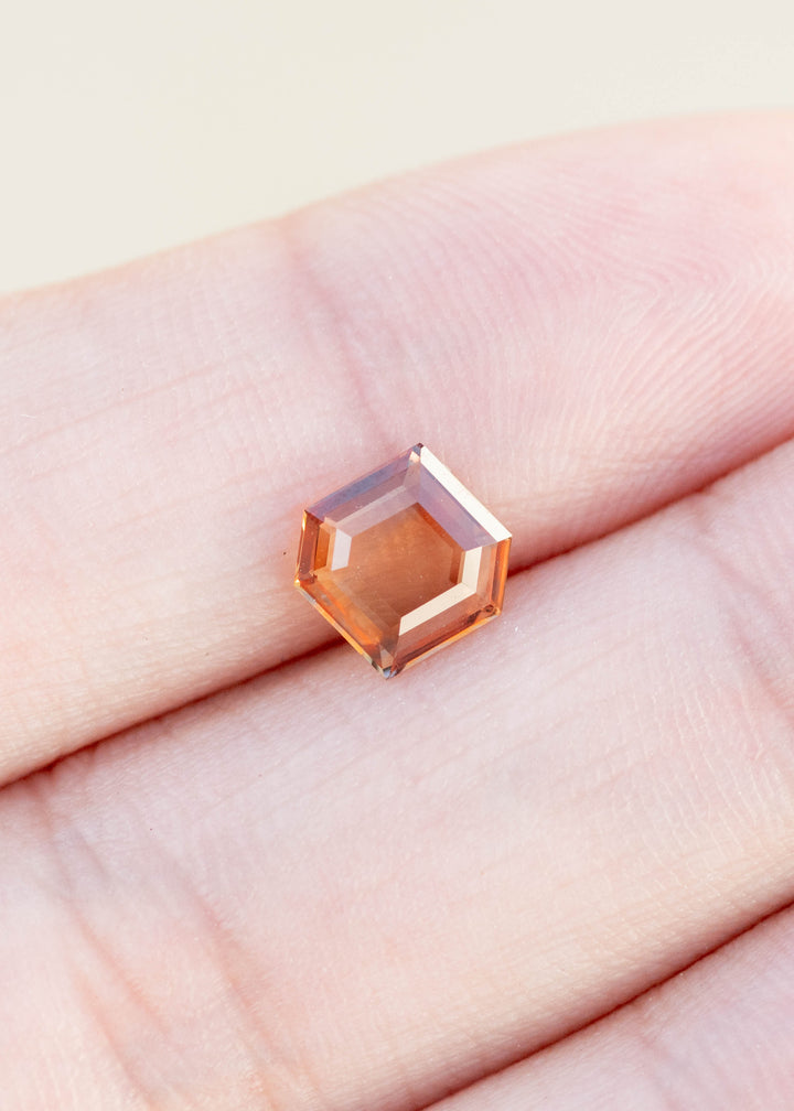 Hexagonal portrait cut sapphire in bright orange held in fingertips under natural light.
