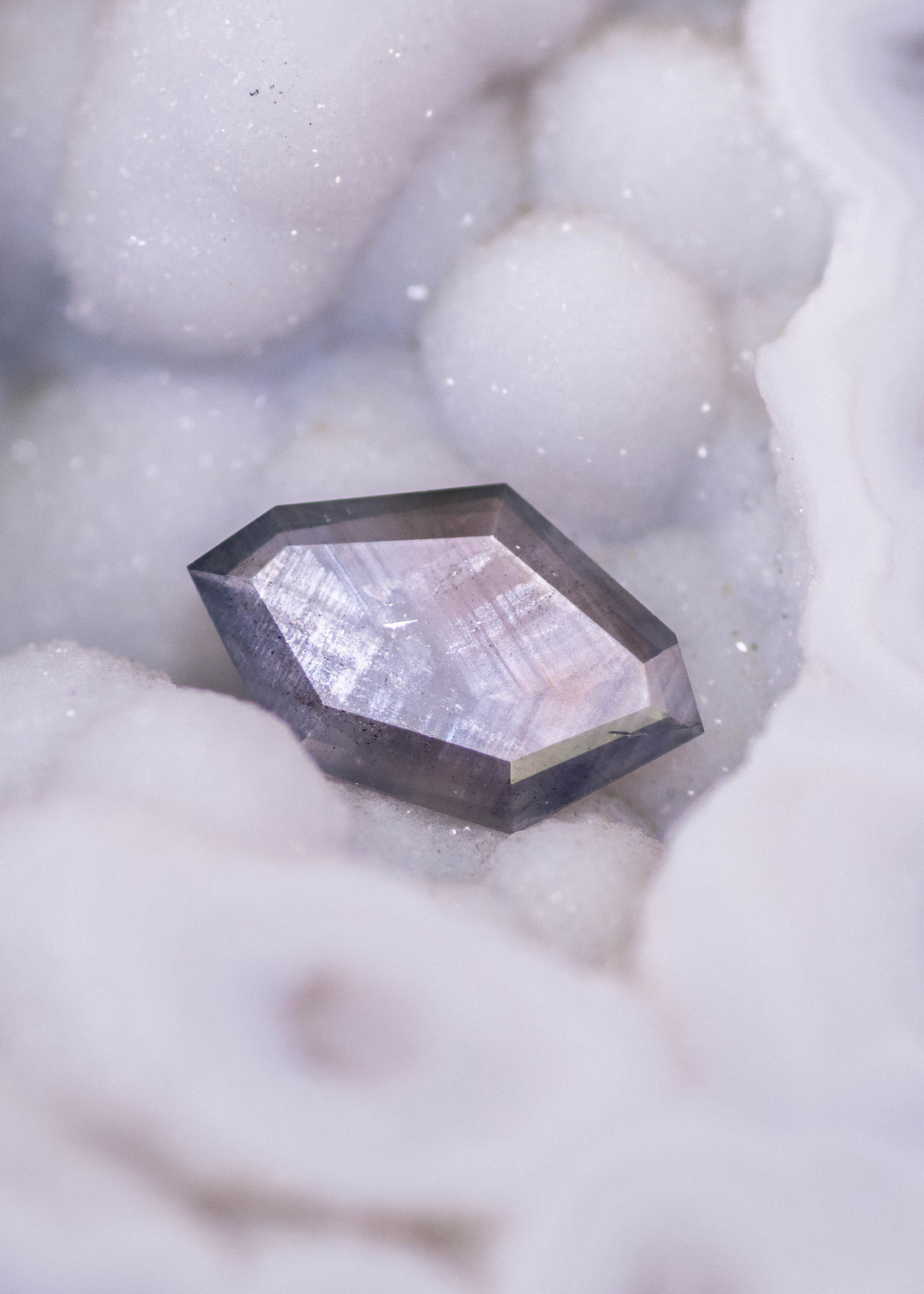 Geometric shield portrait cut sapphire photographed resting in white mineral matrix, showing purple brown gray parti color zoning in an untreated 1.22ct Sri Lanka sapphire.