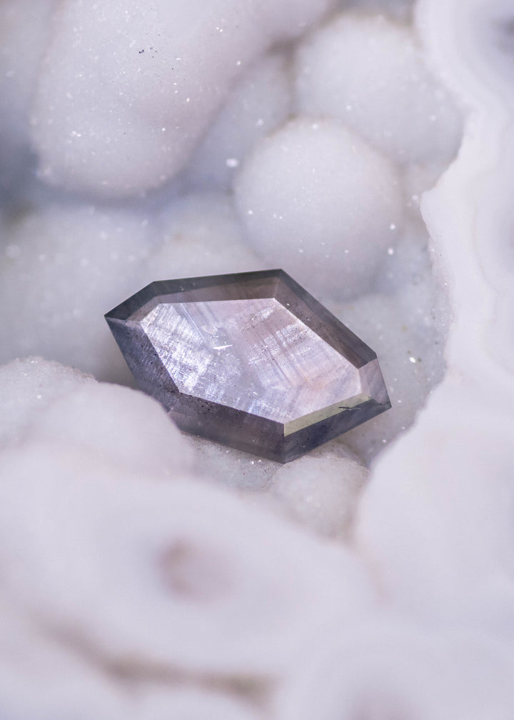 Geometric shield portrait cut sapphire photographed resting in white mineral matrix, showing purple brown gray parti color zoning in an untreated 1.22ct Sri Lanka sapphire.