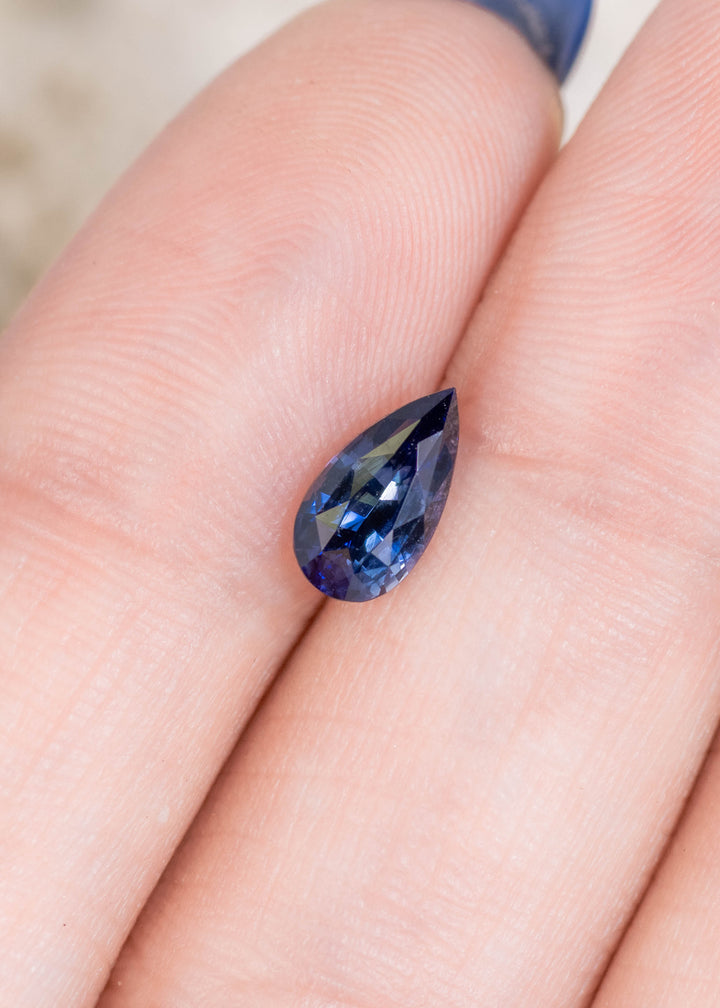 Loose denim blue sapphire from Sakaraha Madagascar shown on a fingertip highlighting the pear shape and bright reflections across the facets.