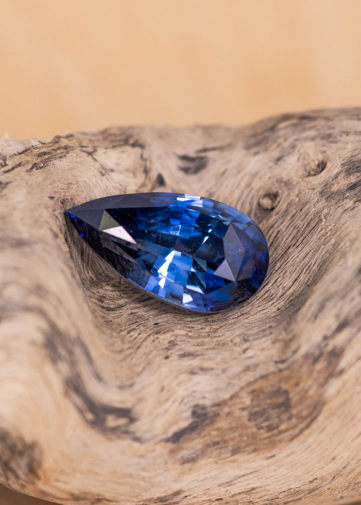 Loose denim blue sapphire from Sakaraha Madagascar resting on natural wood showing the mixed brilliant pear cut and deep blue color.