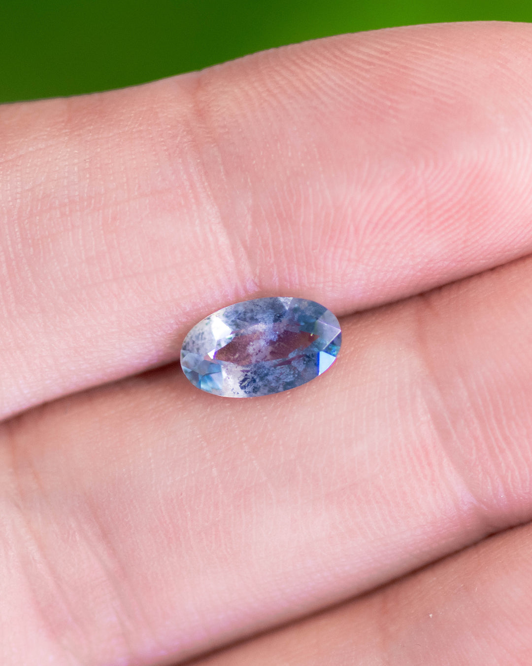 1.24 carat gradient steely blue sapphire resting on a fingertip under natural light, showing elongated oval cut facets and internal inclusions that highlight its natural origin from Umba River Valley, Tanga, Tanzania.