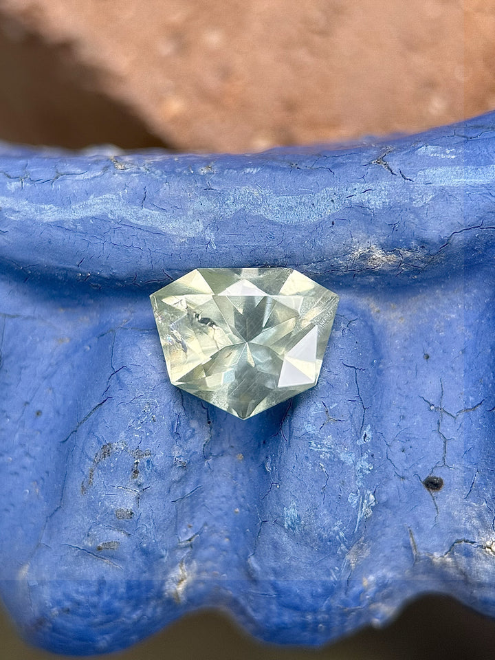 Shield brilliant cut sapphire from Chimwadzulu Hill, Malawi, in yellow-green-blue hues, resting on a weathered royal blue ceramic surface with a terra-cotta background.