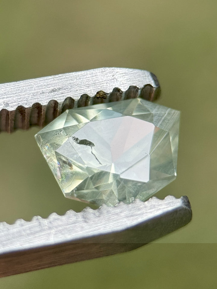 Macro detail of untreated shield brilliant cut sapphire gripped in silver tweezers, revealing internal characteristics and color zoning in green, yellow, and blue against an earthy outdoor background.