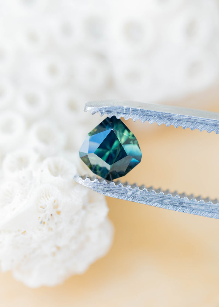 Angular pear-shaped sapphire held between silver tweezers against a softly blurred cream background. Its dark blue-green center is framed by lighter teal and blue reflections, with natural internal features visible through the facets.