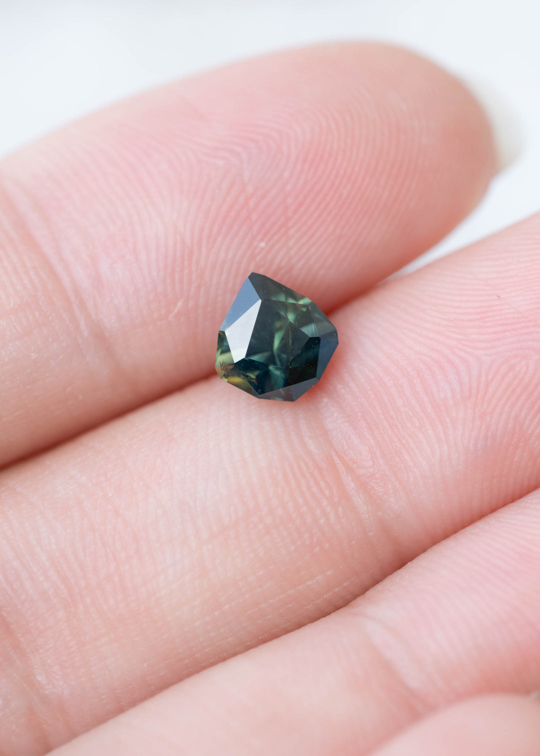 Small, angular blue-green sapphire resting between two fingertips. The gemstone has a dark teal center, broad geometric facets and flashes of lighter green and blue near its edges.
