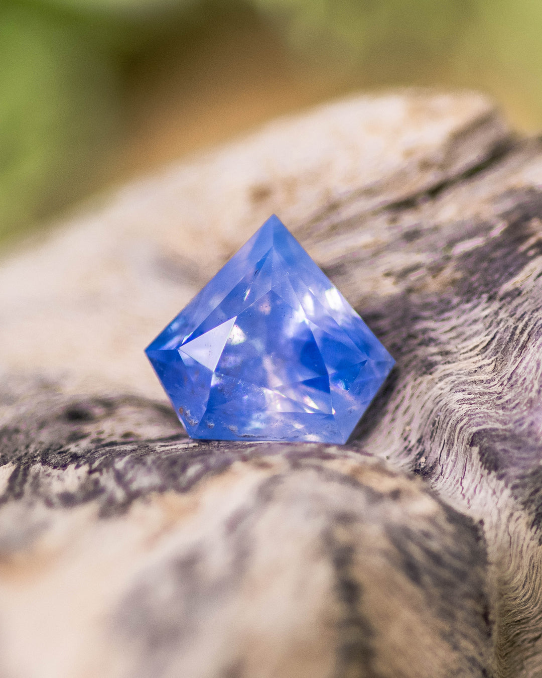 1.28 carat cornflower blue parti sapphire displayed on natural wood, revealing opalescent reflections and fine faceting in shield modified brilliant cut, untreated sapphire from Sakaraha, Madagascar.