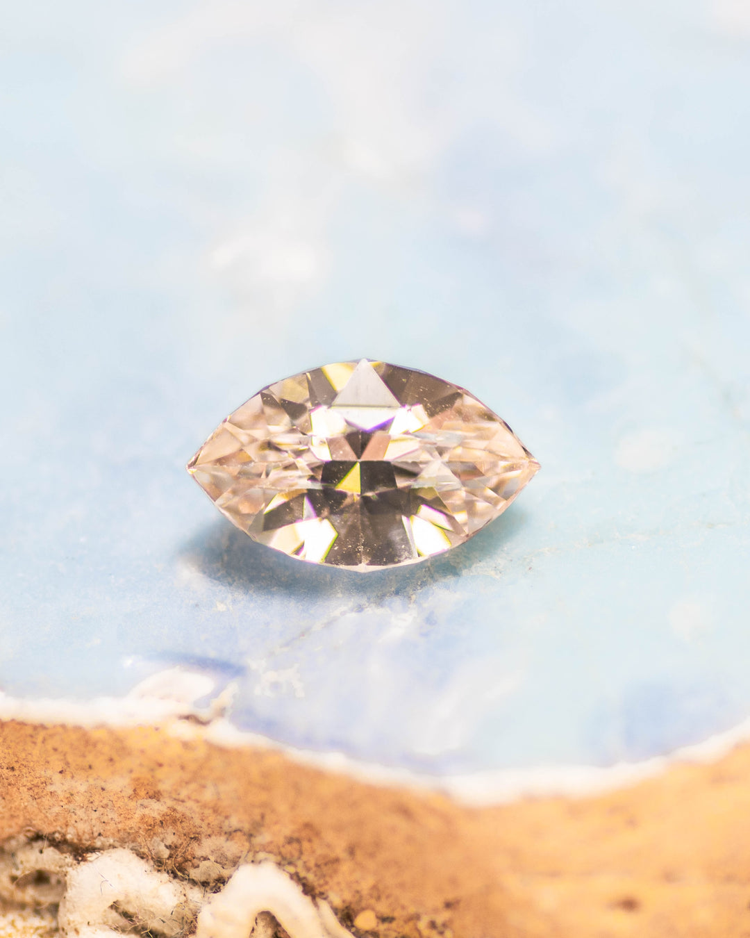 1.28 carat peach sapphire resting on a light blue stone background, showing its warm pastel hue and brilliance from the precision marquise mixed cut, untreated sapphire from Sakaraha, Madagascar.