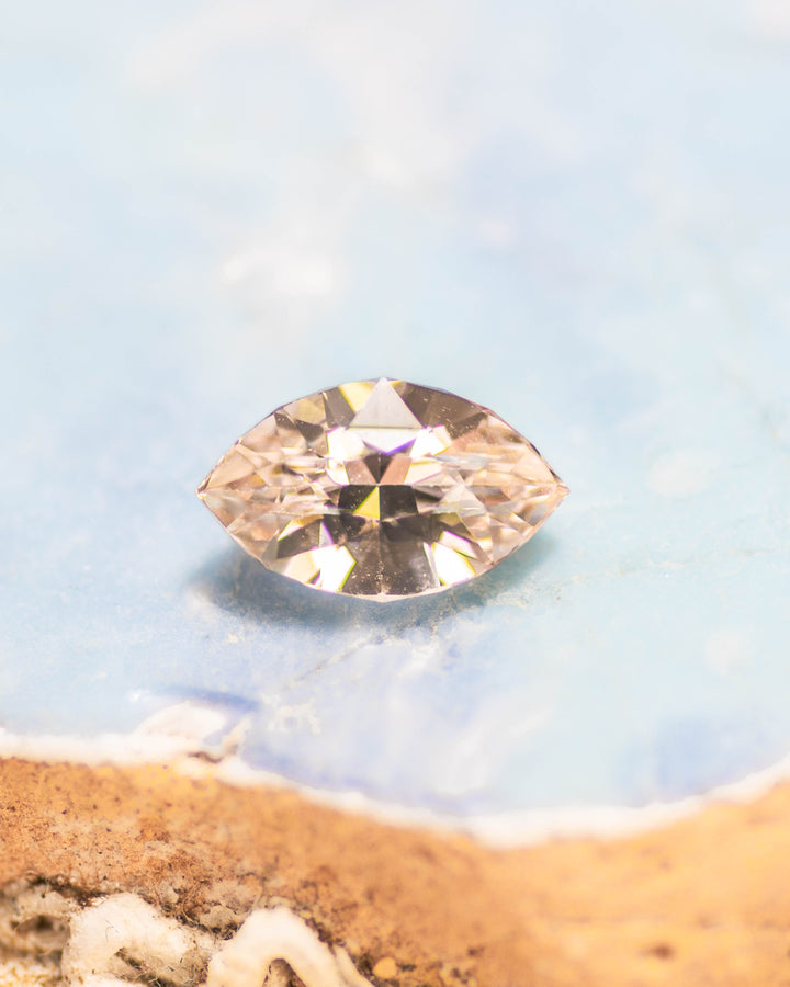 1.28 carat peach sapphire resting on a light blue stone background, showing its warm pastel hue and brilliance from the precision marquise mixed cut, untreated sapphire from Sakaraha, Madagascar.