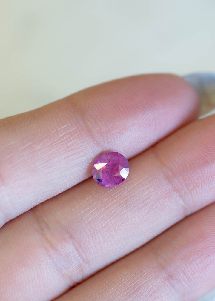 Oval mix cut opalescent pink sapphire resting on fingertips, showing vivid rose-pink tones and a subtle blue color zone near the edge of the stone.