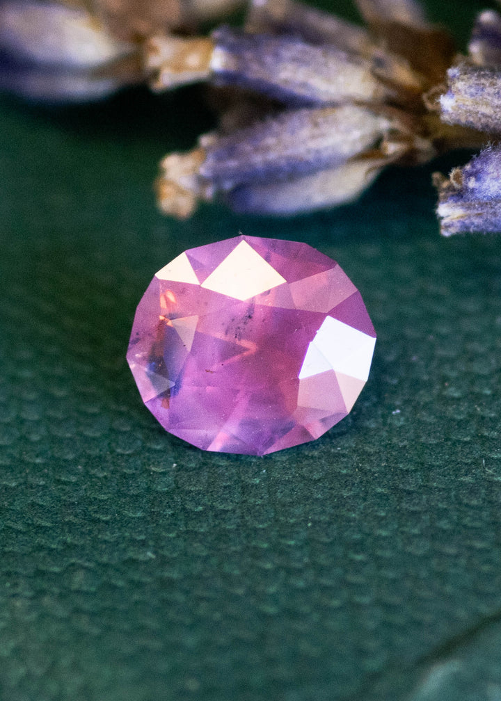 Pink opalescent sapphire on a textured green surface with blurred lavender flowers in the background