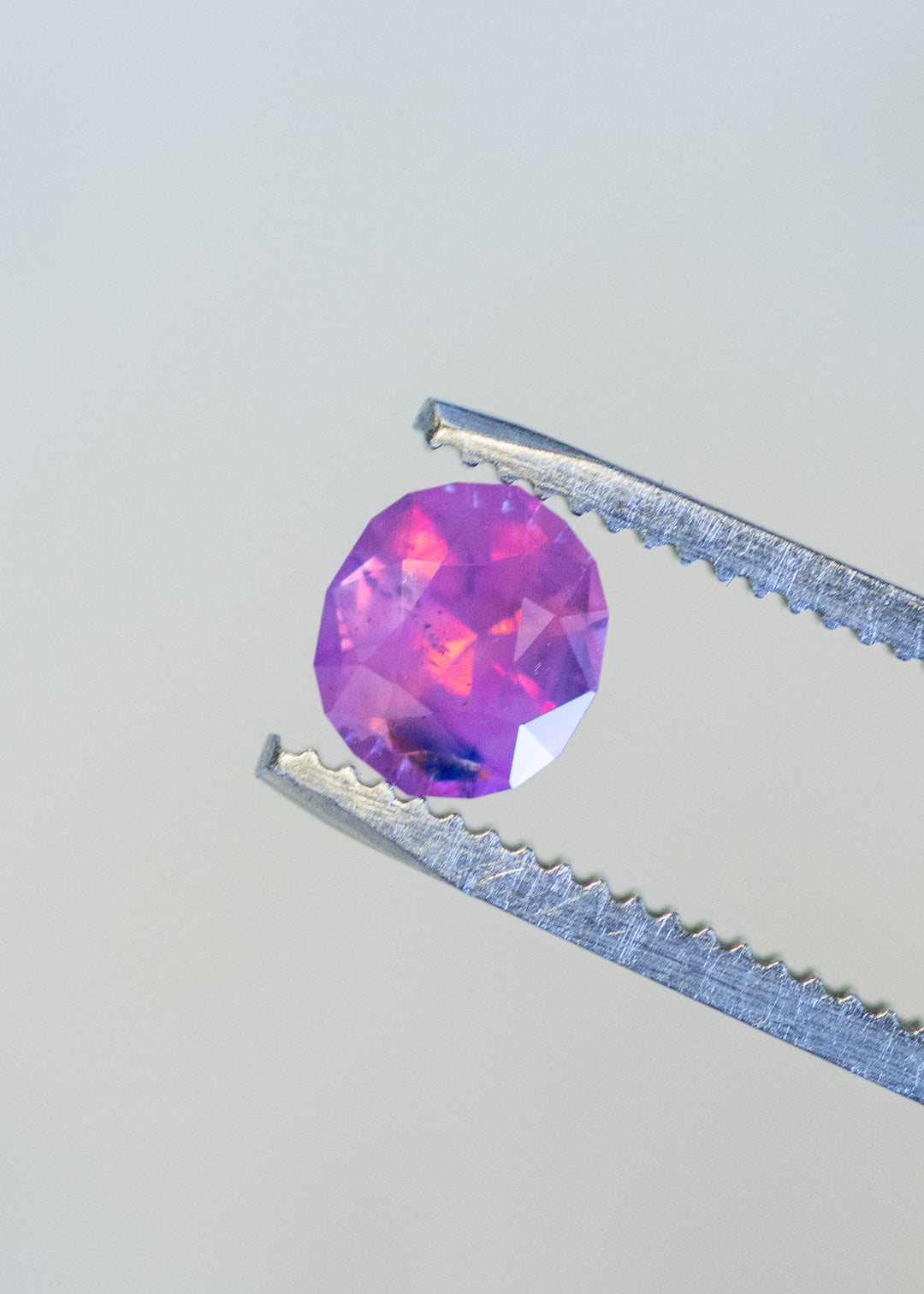 Natural unheated pink sapphire with blue parti zoning held in tweezers, highlighting oval mix cut brilliance and vibrant pink color saturation.