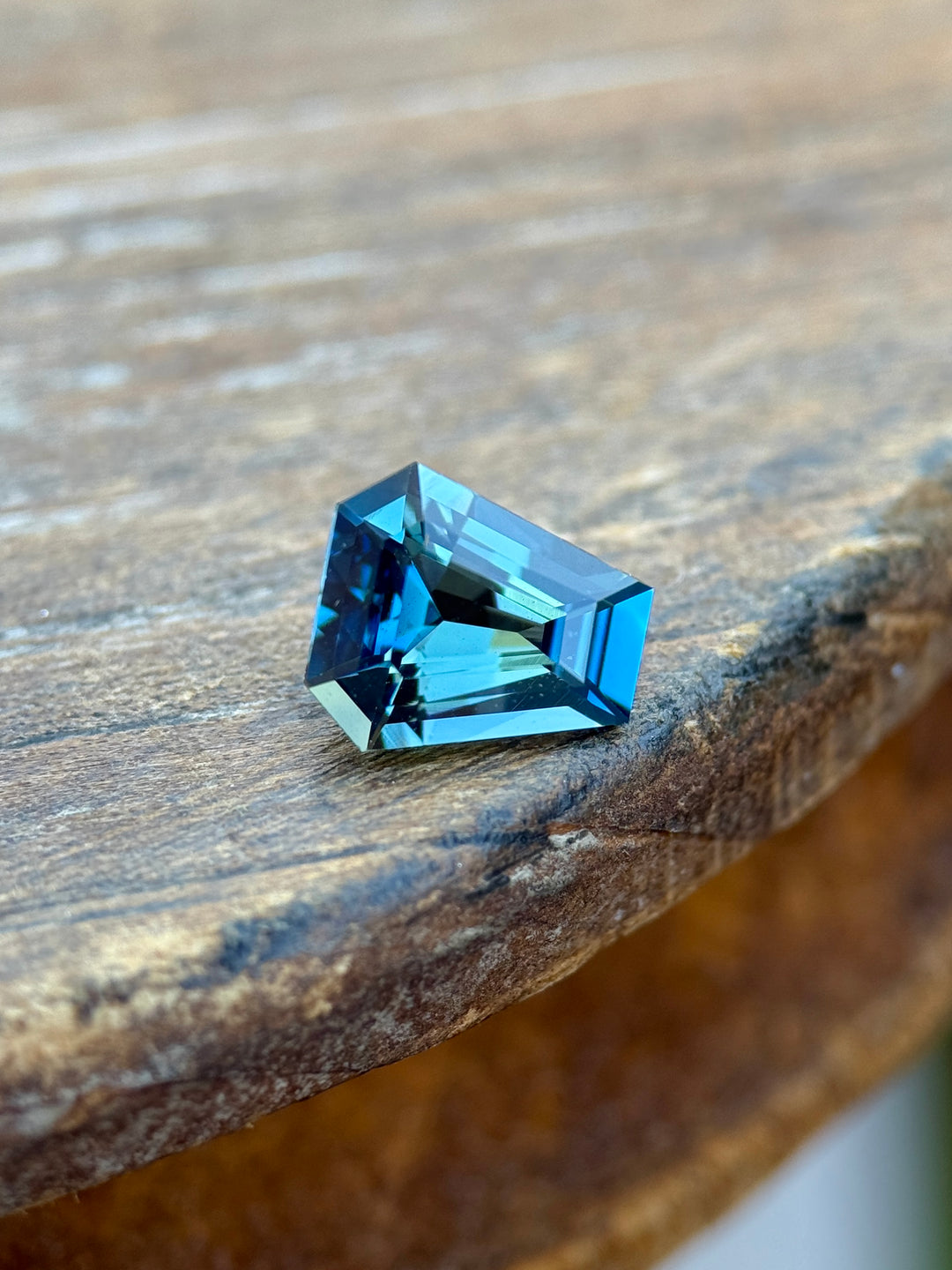 Vibrant 1.29 carat greenish teal blue sapphire from Diego, Madagascar resting on a wooden surface. The custom coffin step cut gemstone showcases clean symmetry, brilliant teal-blue reflections, and natural untreated clarity.