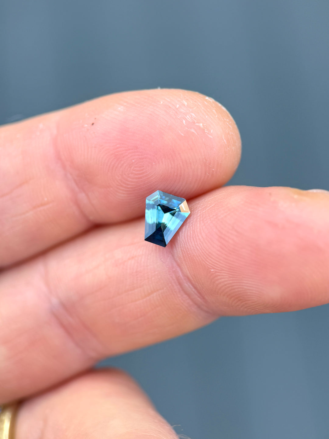 Macro shot of 1.29 carat vibrant greenish teal blue sapphire from Diego, Madagascar balanced on fingertips. The untreated coffin step cut gem displays rich teal tones, precision craftsmanship, and exceptional brilliance.