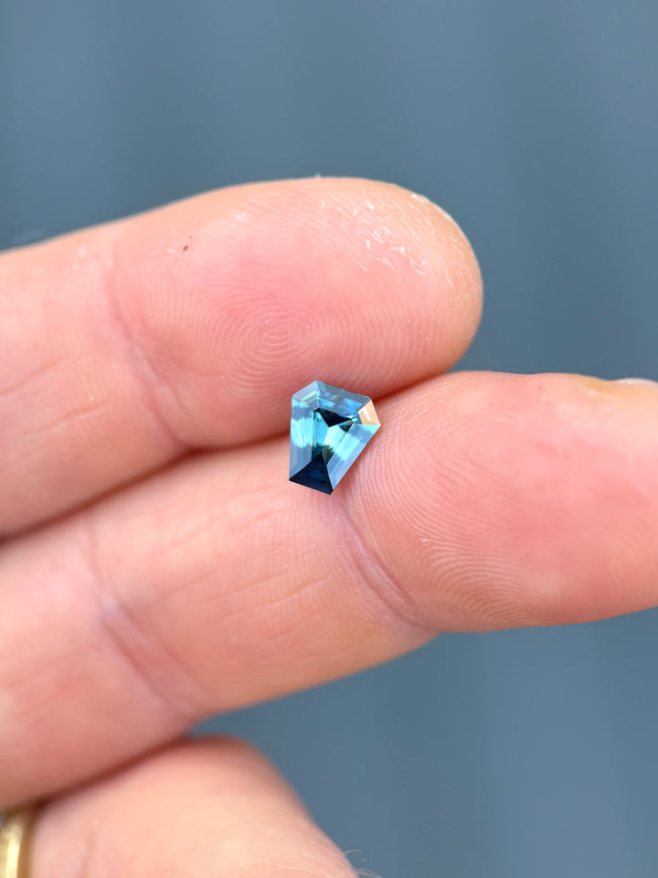 Macro shot of 1.29 carat vibrant greenish teal blue sapphire from Diego, Madagascar balanced on fingertips. The untreated coffin step cut gem displays rich teal tones, precision craftsmanship, and exceptional brilliance.