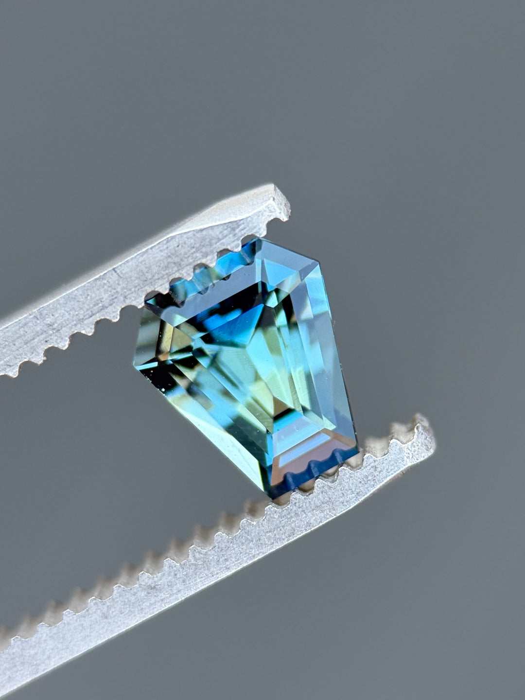 Close-up of 1.29 carat greenish teal blue sapphire from Diego, Madagascar held in gem tweezers. The custom coffin step cut reveals geometric facets, vivid color zoning, and mirror-like luster under natural light.