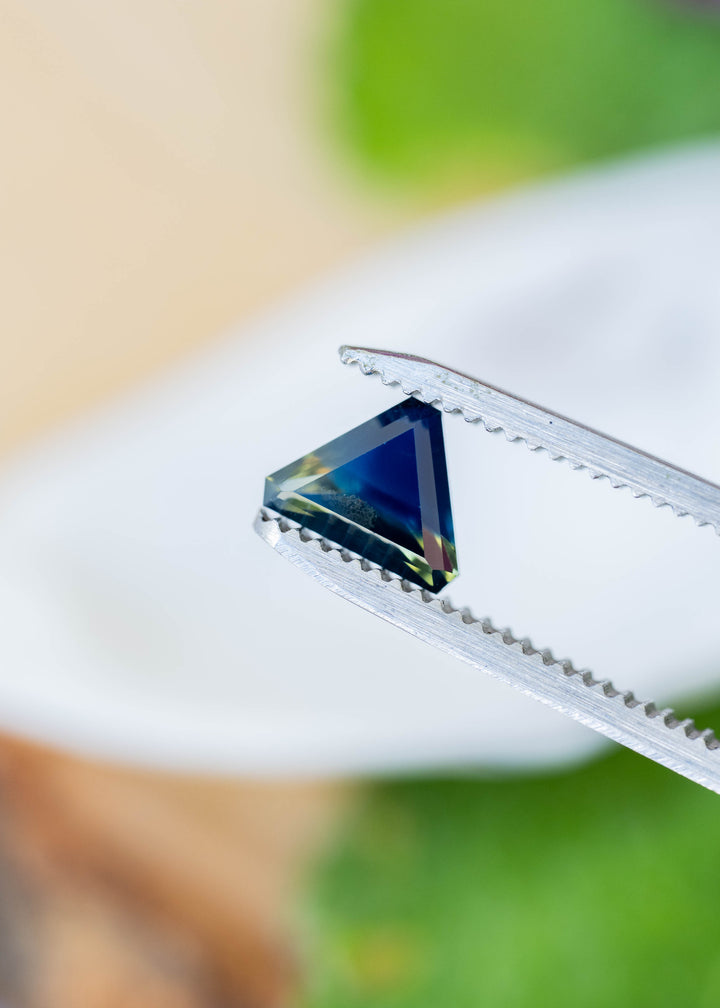 Deep blue triangular sapphire held between silver tweezers against a softly blurred white and green background. Yellow-green areas appear along the lower edge and one corner.