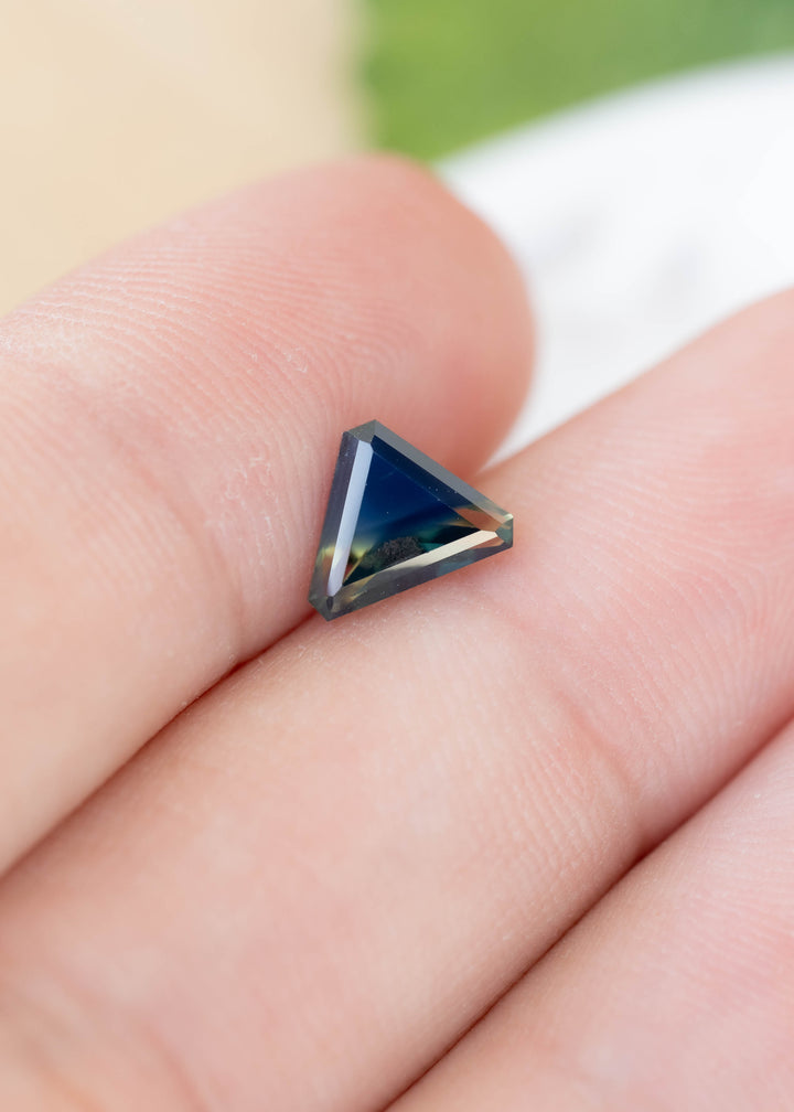 Small triangular sapphire resting across two fingertips. Its center appears dark blue, with a pale yellow-green band along the lower edge and crisp, angular facets.