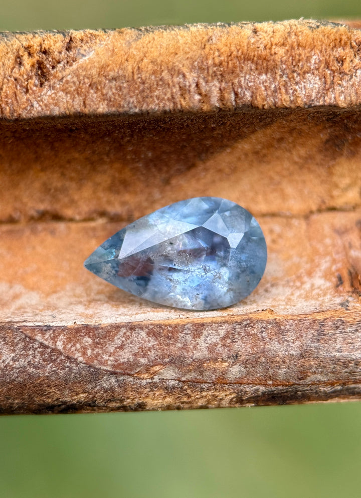 1.30 carat periwinkle blue sapphire resting on textured wood, displaying natural inclusions and soft blue hues in a pear mix cut, untreated and ethically sourced from the Umba River Valley, Tanga, Tanzania.